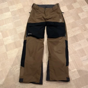 Bruna och svarta skidbyxor Gor Tex från Haglöfs - Säljer ett par tåliga skidbyxor från Haglöfs i brunt och svart. Byxorna är tillverkade i Gore-Tex-material och har praktiska dragkedjor och knappar. Perfekta för utomhusaktiviteter med justerbar midja och förstärkta partier. Byxor har även snöfång som nya.