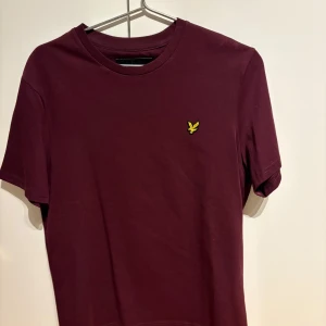 Vinröd t-shirt från Lyle & Scott - Snygg vinröd t-shirt från Lyle & Scott med deras ikoniska logga broderad på bröstet. Perfekt för en stilren och avslappnad look. Tillverkad i mjukt material för extra komfort.