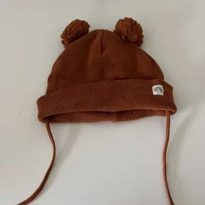 Brun baby mössa med tofsar från Lindex - Söt brun mössa för baby från Lindex med två tofsar på toppen och knytband. Tillverkad i mjukt material med en liten regnbågsdetalj på sidan. Perfekt för att hålla de små varma under kyliga dagar.