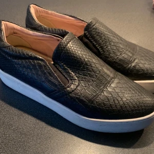 Snygga sneakers från XIT  - Snygga svarta slip-on sneakers med ett coolt ormskinnsmönster. Skorna har en vit sula och elastiska paneler för enkel på- och avtagning. Perfekta för en stilren look. Mycket fint skick. Storlek 39