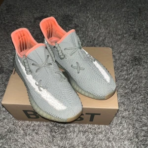 Yeezy Boost 350 - Snygga Adidas Yeezy Boost 350 V2 i grått med orange insida. Skorna har en stickad ovandel och en genomskinlig sula med Boost-dämpning. Perfekta för en trendig look.