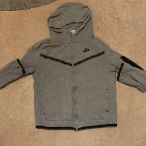 Grå huvtröja från Nike - Säljer en grå huvtröja från Nike med dragkedja och svarta detaljer. Tröjan har en justerbar huva och långa ärmar. Perfekt för en sportig look.