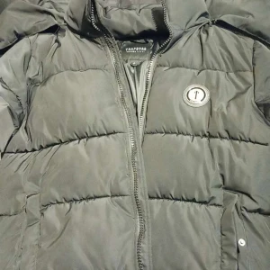 Svart pufferjacka från Trapstar - Snygg svart pufferjacka från Trapstar med huva och dragkedja. Jackan har ett stort Trapstar-tryck i rött på ryggen och en rund logotyp på bröstet. Perfekt för kyliga dagar. priset kan diskuteras vid snabb affär 