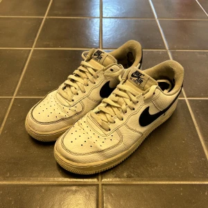 Nike Air Force 1 i vit och svart - Säljer ett par klassiska Nike Air Force 1 sneakers i vit med svarta detaljer. Skorna har snörning och en robust sula. Perfekta för en stilren look.