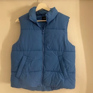 Blå dunväst från Lindex - Säljer en snygg blå dunväst från Lindex med dragkedja framtill. Västen har en hög krage och två praktiska fickor. Perfekt för kyliga dagar!