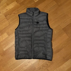 Grå dunväst från J.Lindeberg - Snygg grå dunväst från J.Lindeberg i storlek S med dragkedja framtill. Perfekt för lager-på-lager under kyliga dagar. Västen har en stilren design med hög krage och är lätt att matcha med olika outfits.