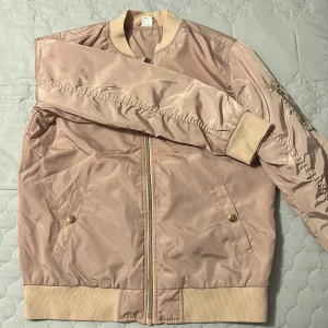 Rosa bomberjacka - Snygg beige bomberjacka med dragkedja och ribbade muddar. Jackan har två fickor med knappar och en klassisk bomberkrage. Perfekt för en stilren look.