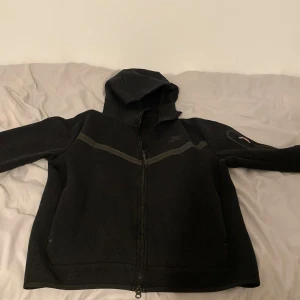 Svart Nike Tech Fleece - Säljer en svart fleecejacka från Nike med huva och dragkedja. Perfekt för kyliga dagar med sin mjuka och bekväma design. Jackan har långa ärmar och en diskret logga på bröstet.