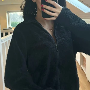 Svart fleecejacka med dragkedja - Mysig svart fleecejacka med dragkedja framtill. Perfekt för kyliga dagar med sin mjuka och varma känsla. Jackan har långa ärmar och en hög krage för extra värme.