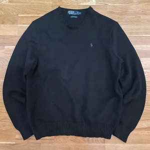 Svart tröja från Polo Ralph Lauren - Säljer en klassisk svart tröja från Polo Ralph Lauren i 100% Pima bomull. Tröjan har en broderad lila logotyp på bröstet och ribbade muddar vid ärmslut och nederkant. Perfekt för en stilren look.