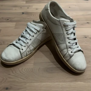 Vita sneakers från Saint Laurent - Snygga vita sneakers från Saint Laurent med klassisk snörning och perforerade detaljer på sidorna. Perfekta för en stilren look. Använda men inget som är sönder med de. Behövs bara en trasa och det är helt redo för att spränga stan 😝