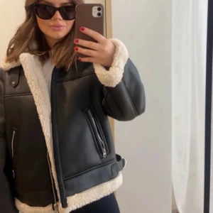 Svart skinnjacka med pälsfoder - Snygg svart skinnjacka med mjukt vitt pälsfoder. Jackan har dragkedja och två fickor med dragkedjor. Perfekt för kyligare dagar med stil.