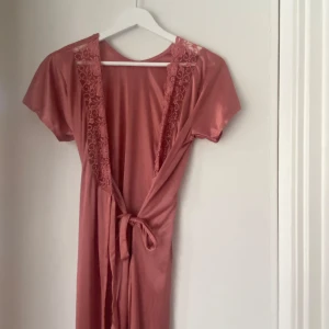 Rosa kimono med spetsdetaljer - Elegant rosa kimono med spetsdetaljer längs halsringningen och ett knytband i midjan. Kimonon har korta ärmar och är tillverkad i ett mjukt, glansigt material som ger en lyxig känsla.