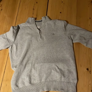 Grå tröja med dragkedja från GANT - Säljer en stilren grå tröja från GANT med dragkedja och ribbade detaljer. Tröjan har en liten broderad logga på bröstet och en randig krage. Perfekt för en avslappnad stil. Storleken är 170 och passar 14-15 åringar. Priset är inte hugget i sten!