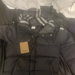 Svart dunjacka från Burberry - Snygg svart dunjacka från Burberry med klassiskt rutigt mönster på insidan av huvan. Jackan har dragkedja och knappar framtill samt en stilren design med Burberry-logga på ärmen.