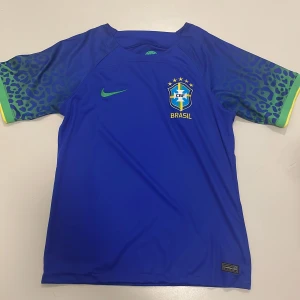 Blå Brasilien fotbollströja från Nike - Snygg blå fotbollströja från Nike med Brasilien-emblem på bröstet. Tröjan har korta ärmar med ett coolt mönster i grönt och gult. Perfekt för fotbollsfans som vill visa sitt stöd för Brasilien! Tröjan är köpt i Brazilen. Skicket är 10/10, använd en gång. De är bara att fråga om ni har några frågor.