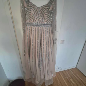 Champagnefärgad långklänning med glittriga detaljer - Elegant champagnefärgad långklänning med glittriga silverdetaljer. Klänningen har en v-ringad design och långa ärmar i ett transparent material. Perfekt för en glamorös kväll.