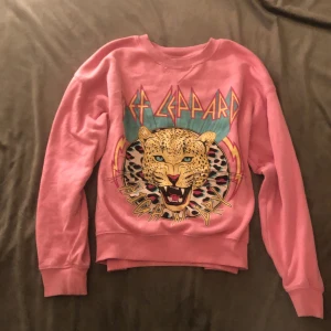 Rosa Def Leppard sweatshirt pink - Säljer en cool rosa sweatshirt med Def Leppard-tryck. Tröjan har ett stort leopardmotiv och färgglada detaljer på framsidan. Perfekt för fans av bandet eller för en retro look. Mjuk och bekväm med ribbad krage och ärmslut🫶🏼