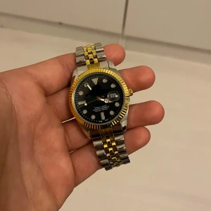 Tvåfärgad klocka med svart urtavla - Snygg klocka med en kombination av silver och guld på armbandet och en svart urtavla. Klockan har en datumvisare och en räfflad guldfärgad bezel. Perfekt för den som gillar en klassisk och stilren look.