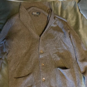 Grå kofta från Polo Ralph Lauren - Säljer en elegant grå kofta från Polo Ralph Lauren, tillverkad i en lyxig blandning av kashmir och ull. Koftan har knappar framtill och en klassisk design som passar perfekt för kyligare dagar.