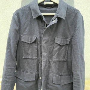 Fieldjacket john Henric - Säljer en marinblå fieldjacket från John Henric i en blandning av linne och bomull. Linnet ligger i fodret på jackan. Jackan har långa ärmar och praktiska fickor framtill. Perfekt för en stilren look. Jackan är använd ett par gånger men den är så gott som ny