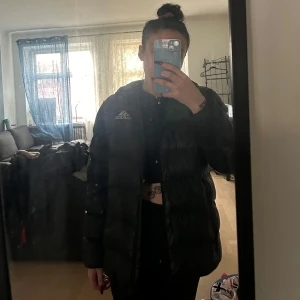 Svart dunjacka från Adidas - Säljer en svart dunjacka från Adidas med huva och dragkedja. Jackan har ett stilrent quiltat mönster och Adidas-loggan på bröstet. Perfekt för kyliga dagar. Passar även XS/S