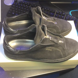 Svarta sneakers från Axel Arigato - Snygga svarta sneakers från Axel Arigato zip i mocka med dragkedja och diskret logga på sidan. Perfekta för en stilren look. 