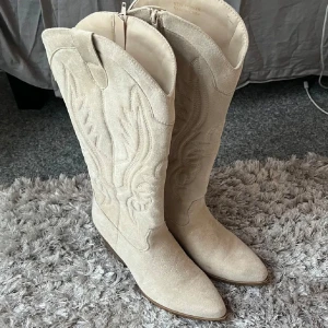 Cowboyboots  - Säljer ett par cowboyboots i storlek 41, skorna är använda 2 ggr. Skorna står endast hemma, de är som nya! 💕💕 hör av dig så kan jag skicka fler bilder.      - Plicks beskrivning: Snygga beige cowboy boots i mocka med broderade detaljer och spetsig tå. De har en dragkedja på sidan för enkel på- och avtagning. Perfekta för en stilren look.