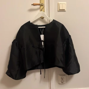 Svart croppad jacka/blus från Gina Tricot - Snygg svart croppad jacka från Gina Tricot med knytband framtill. Jackan har en elegant och glansig finish, perfekt för en stilren look. Den har vida ärmar som ger en trendig touch. Perfekt till jul eller nyår. Oanvänd med lappen kvar. Inköpt för 499.95 kr.