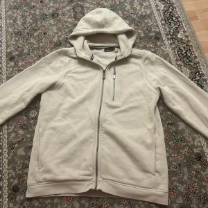 Sail racing kofta med huva - Säljer en kofta från sail racing med huva och dragkedja framtill. Koftan har långa ärmar och en bröstficka med dragkedja. Perfekt för kyliga dagar!