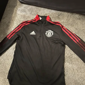 Svart Manchester United tröja från Adidas - Säljer en svart långärmad Manchester United tröja från Adidas med röda detaljer och dragkedja vid halsen. Perfekt för fotbollsfans! Tröjan har klubbens emblem på bröstet och är tillverkad i Primegreen-material.
