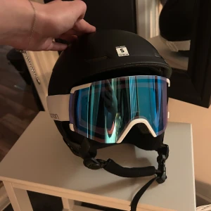 Skidhjälm med skidglasögon  - Säljer en svart hjälm i storlek S tillsammans med blå skidglasögon från Salomon. Helt ny och oanvänd. Nytt pris ca 2000