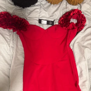 Röd långklänning med blommiga detaljer - Elegant röd långklänning med blommiga detaljer på axlarna. Klänningen har en figurnära passform och en vacker, svepande kjol. Perfekt för speciella tillfällen. Klänningen har aldrig använts råkat bestämma i fel storlek. 