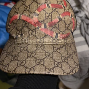 gucci keps Beige med ormmönster  - Snygg beige keps från Gucci med ett iögonfallande ormmönster i rött och svart. Kepsen har det klassiska Gucci-mönstret i bakgrunden och är tillverkad i Italien. Perfekt för att ge din outfit en lyxig touch.