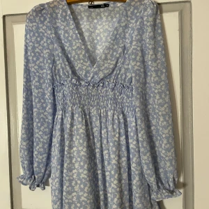 Blå blommig klänning från 157 - Charmig blå klänning med vita blommor från 157. Den har en v-ringad design och långa ärmar med elastisk midja för en bekväm passform. Perfekt för vår och sommar!