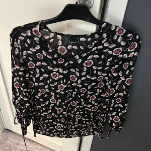 Blommig blus från Gina Tricot - Säljer en söt blommig blus från Gina Tricot i svart med rosa och vita blommor. Blusen har trekvartsärmar med knytning och en rund halsringning. Perfekt för en stilren look! Använd bara en gång och är i mycket bra skick!