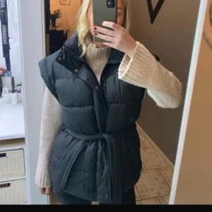 Svart dunväst med bälte - Snygg svart dunväst med bälte i midjan. Västen har en hög krage och knappar framtill, perfekt för kyligare dagar. Den är stilren och lätt att matcha med olika outfits. Det är smink fläckar p kragen.