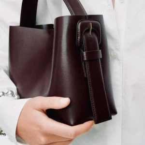 Brun axelremsväska från ARKET - Snygg brun handväska i skinn från arket  med stilren design. Väskan har ett brett handtag och en elegant spänndetalj och axelrem . Perfekt för att ge en touch av lyx till din outfit. Detta är mellan storlek.. helt ny och oanvänd!