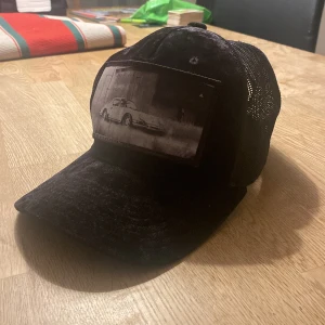 Svart keps med bilmotiv från Hatstore - Cool svart keps med ett retro bilmotiv på framsidan. Kepsen har ett nätmaterial baktill för extra ventilation och en justerbar rem för perfekt passform. Perfekt för en avslappnad stil.