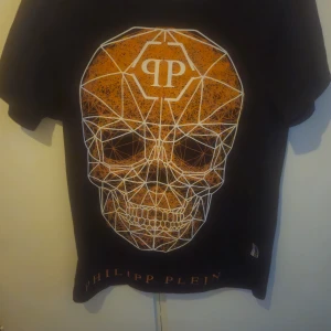 Svart t-shirt med dödskalle från Philipp Plein - Cool svart t-shirt från Philipp Plein med ett stort geometriskt dödskalletryck i orange och vitt på framsidan. T-shirten har korta ärmar och en klassisk passform. Perfekt för dig som vill sticka ut med en edgy stil. kvitto finns