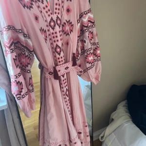 Buda på NY Rosa klänning från Indiska - Säljer en vacker rosa klänning från Indiska med ett etniskt mönster i olika rosa nyanser. Klänningen har långa ärmar och ett knytbälte i midjan för en smickrande passform. Perfekt för en bohemisk look!