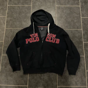 Svart hoodie från Vinson Polo Club - Säljer en svart hoodie från Vinson Polo Club med röd text på bröstet. Den har en dragkedja framtill och en mysig huva med snören. Perfekt för en skön och clean look. Storlek L men sitter mer som en S/M.