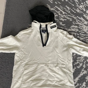 Vit hoodie från Woolrich - Säljer en stilren vit hoodie från Woolrich med svart dragkedja och en praktisk ficka framtill. Huva är infällbara och göms då snyggt i kragen. 