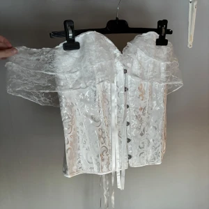 Vit spetskorsett med snörning - Vit offshoulder korsett i spets med snörning på baksidan och knappar framtill. Helt oanvänd då jag köpte två. På lappen står det strl L men passar fler storlekar då man spänner den själv i ryggen. Perfekt som festtopp eller nu till jul.