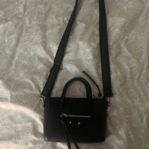 Svart axelväska med dragkedja - Snygg svart axelväska med justerbar rem och praktisk dragkedja framtill. Perfekt för att bära dina dagliga nödvändigheter med stil.