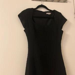 Svart klänning med korta ärmar - Elegant svart klänning med korta ärmar och en fyrkantig halsringning. Klänningen har en figurnära passform och en diskret dragkedja i ryggen.