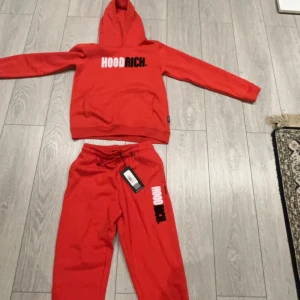 Röd hoodie från Hoodrich - Säljer en cool röd hoodie från Hoodrich med texten 'HOODRICH' i vitt och svart på bröstet. Den har en klassisk känguruficka och en bekväm huva. Perfekt för en avslappnad stil.