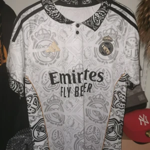 Vit fotbollströja från Adidas - Snygg vit fotbollströja från Adidas med Real Madrid-logga och gulddetaljer. Tröjan har korta ärmar och ett unikt mönster i grått. Perfekt för alla fotbollsfans!