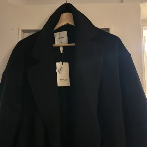 Svart kappa från Object - Snygg svart kappa från Object med klassisk design. Den har långa ärmar och ett bälte i midjan för en elegant look. Perfekt för kyligare dagar.