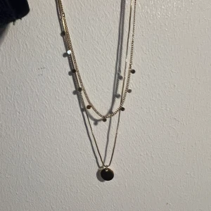 Guldigt halsband med hängsmycke - Elegant guldigt halsband med dubbla kedjor och små runda plattor. Det nedre lagret har ett runt hängsmycke som ger en stilren look. Perfekt för att lägga till en touch av elegans till din outfit.